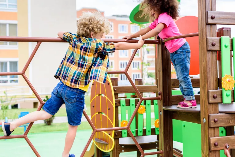 Two children playing on a jungle gym. One child is climbing a hexagonal net, while the other child stands on a platform reaching out. Colorful playground equipment and apartment buildings in the background.